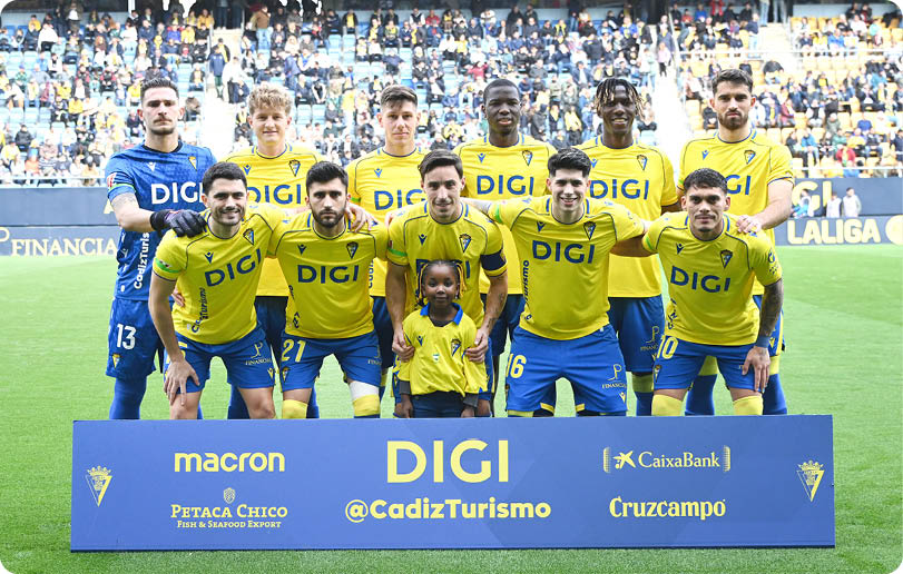 A group of men and a boy pose for a photo on a field. They are wearing yellow and blue shirts and are standing in front of a blue sign. Contenido generado con IA