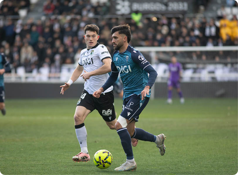 Two soccer players are on the field, one wearing a blue shirt and the other wearing a white shirt. They are both running towards the ball. Contenido generado con IA
