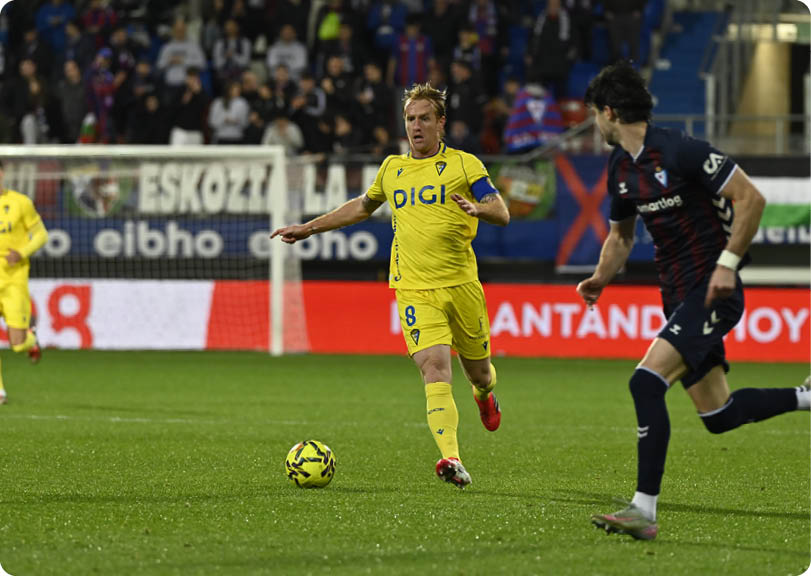 Two soccer players are running towards a ball on a field. One player is wearing a yellow shirt and the other is wearing a blue shirt. Contenido generado con IA