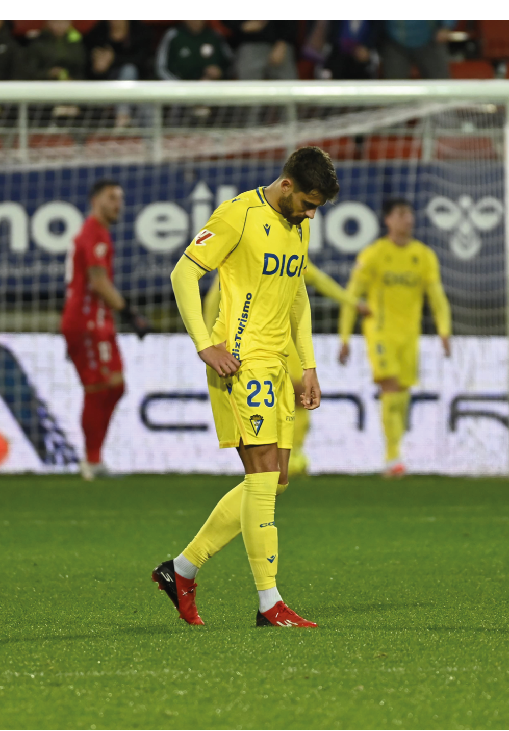 A soccer player in a yellow jersey is walking away from the field, looking down at the ground. Contenido generado con IA