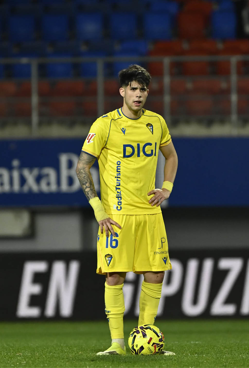 A soccer player in a yellow uniform stands on the field with a soccer ball in front of him. Contenido generado con IA
