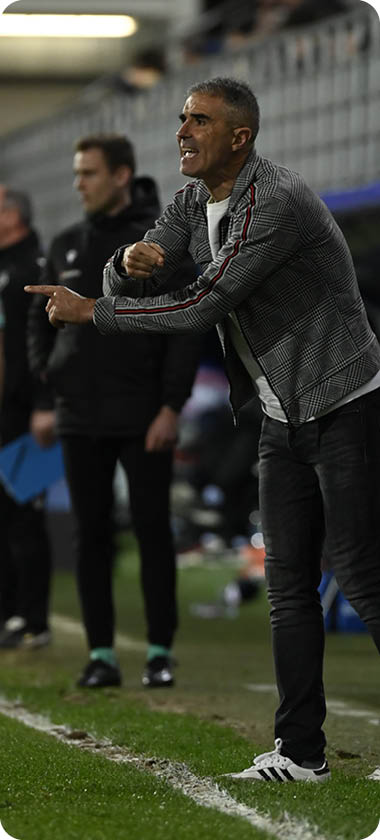 A man in a grey jacket is standing on a soccer field, pointing at something. Contenido generado con IA