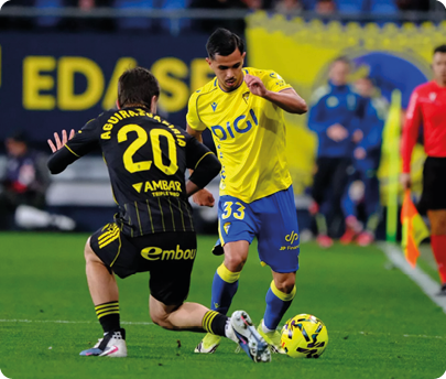 Two soccer players are on the field, one is kicking the ball while the other is trying to block him. Contenido generado con IA