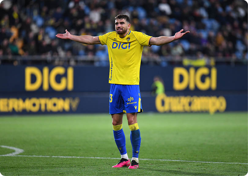 A soccer player in a yellow and blue uniform stands on the field with his arms outstretched. Contenido generado con IA