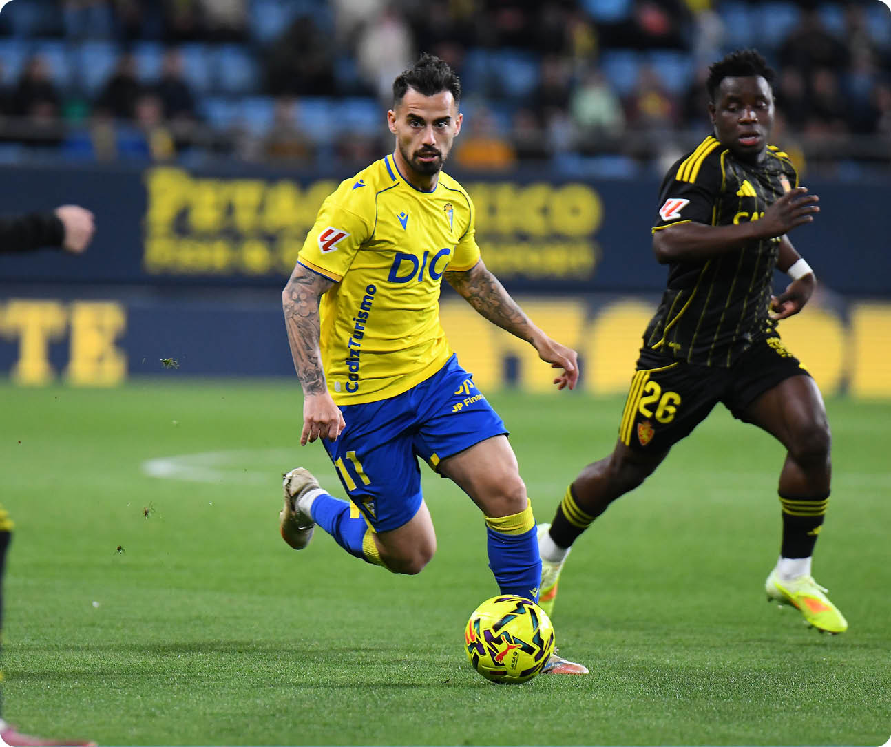 Three soccer players are on the field, one of them wearing a yellow and blue uniform. The player in the center is kicking a soccer ball. Contenido generado con IA
