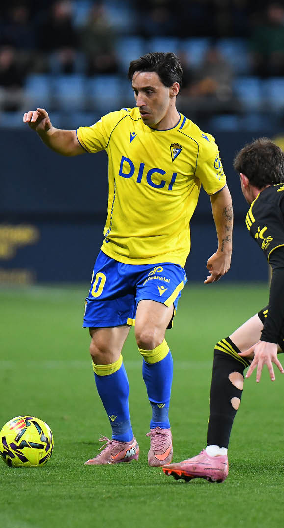 Two soccer players on a field, one wearing a yellow shirt and the other wearing a black shirt. The yellow player is kicking the ball while the black player tries to block him. Contenido generado con IA