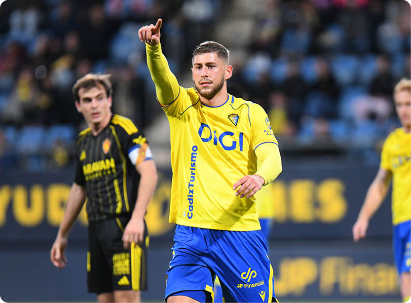 A soccer player in a yellow and blue uniform points to the sky, while his teammates look on. Contenido generado con IA