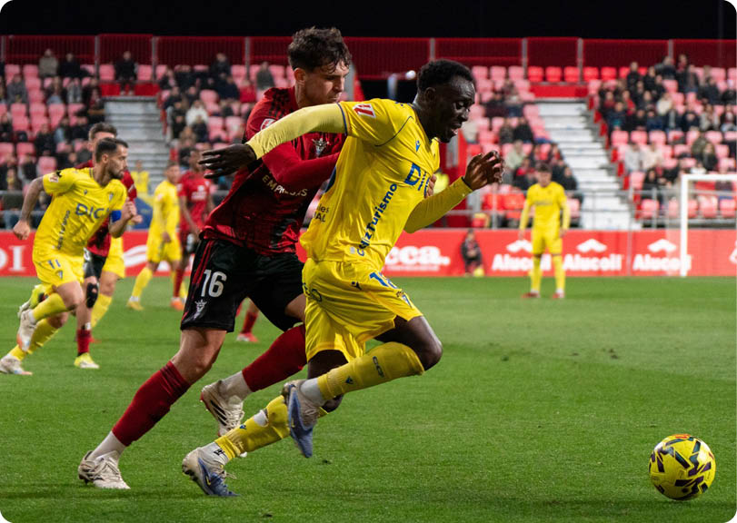 Two soccer players are running for the ball on a field. One player is wearing a yellow shirt and the other is wearing a red shirt. Contenido generado con IA