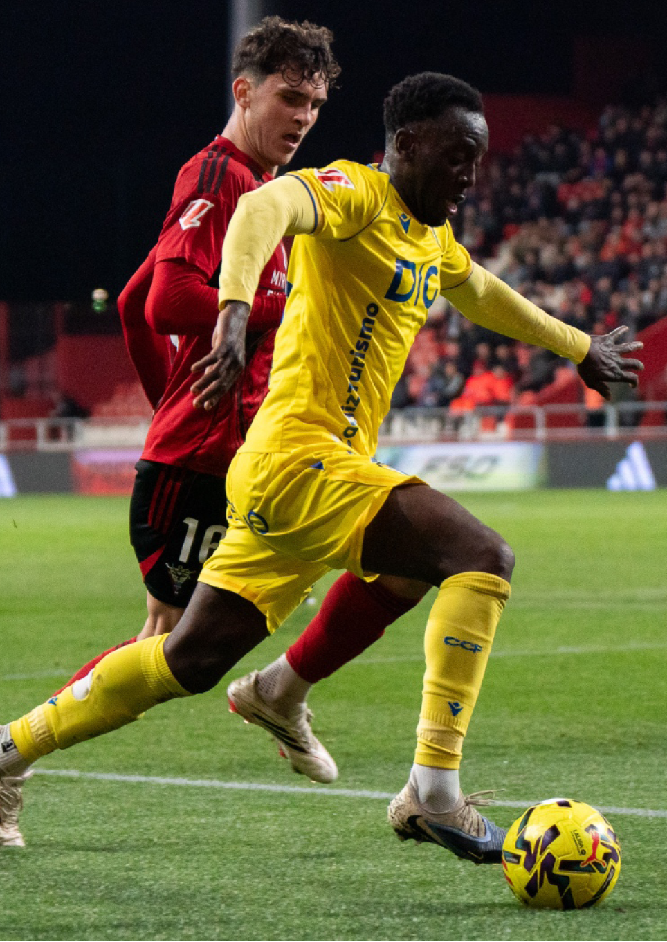 Two soccer players are on the field, one in a yellow shirt and the other in a red shirt. The yellow player is about to kick the ball. Contenido generado con IA