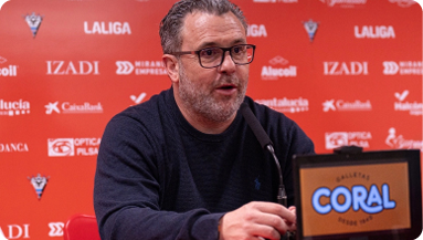 A man in a blue sweater is sitting at a table with a microphone in front of him. Contenido generado con IA