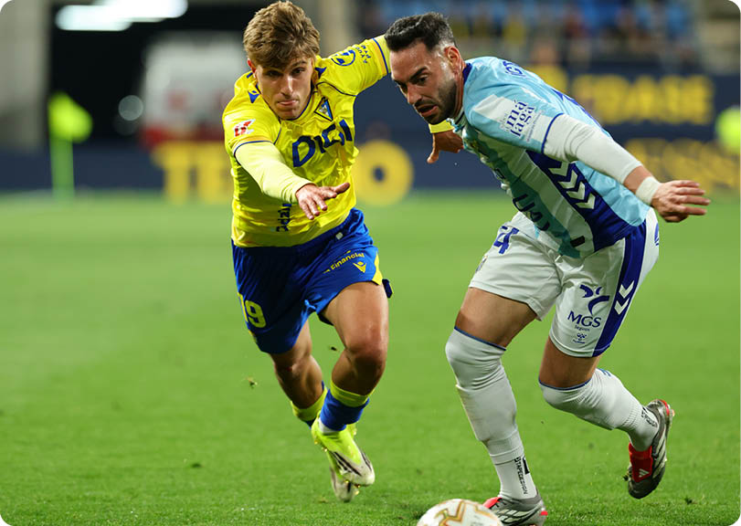 Two soccer players are running for the ball on a field. One player is wearing a yellow shirt and the other is wearing a red shirt. Contenido generado con IA