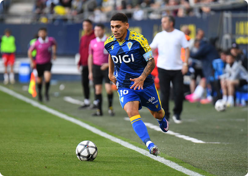 A soccer player in a blue and yellow uniform is running towards a soccer ball. Descripci n generada con IA