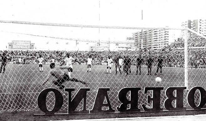 Un jugador est pateando un bal n en un campo de f tbol mientras otros jugadores y personas est n alineados frente a una multitud de espectadores en las gradas.  Descripci n generada con IA