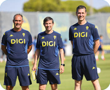 Tres adultos con ropa deportiva azul con el logo DIGI posan sonrientes en un campo de entrenamiento al aire libre. Descripci n generada con IA