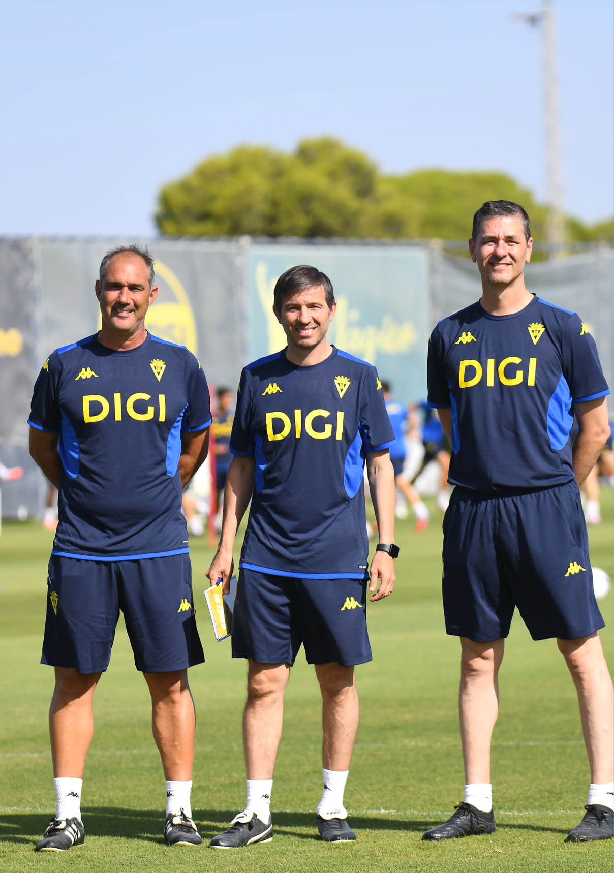 Tres adultos con ropa deportiva azul con el logo DIGI posan sonrientes en un campo de entrenamiento al aire libre con otras personas al fondo. Descripci n generada con IA