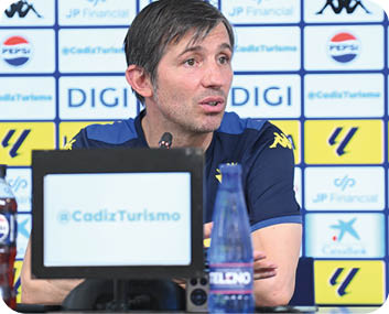 Una persona con camiseta deportiva azul habla en una conferencia de prensa con un micr fono frente a un fondo con logotipos de patrocinadores y una pantalla que muestra el texto @CadizTurismo. Descripci n generada con IA