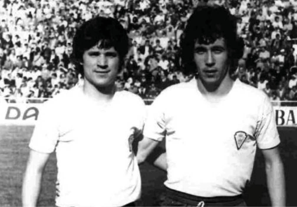Dos personas con camisetas blancas posan juntas en un estadio deportivo con una multitud de fondo. Descripci n generada con IA