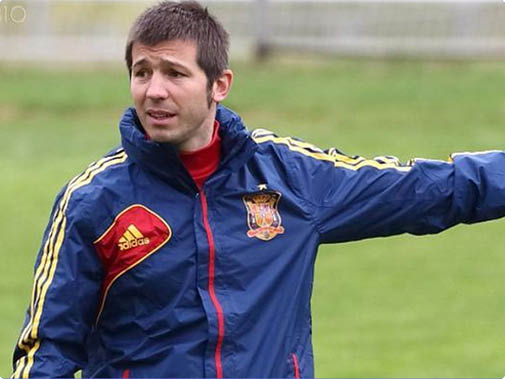 Una persona con chaqueta deportiva azul con detalles en rojo y amarillo se ala hacia un lado en un campo de c sped. Descripci n generada con IA