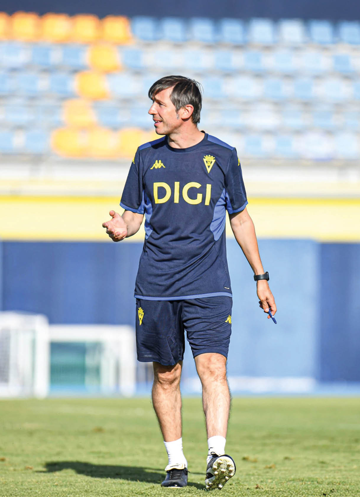 Una persona con ropa deportiva azul oscuro con el logo DIGI camina sobre un campo de c sped en un estadio con gradas vac as al fondo. Descripci n generada con IA