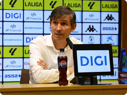 A person in a white shirt is sitting at a table with a microphone and a bottle of Pepsi in front of a backdrop covered with various sponsor logos. Descripci n generada con IA