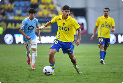 Two people in yellow and blue sports uniforms and one person in a light blue and white uniform are playing soccer on a grass field with one person in yellow controlling the ball. Descripci n generada con IA