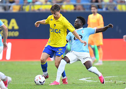 Two people in sports uniforms compete for the soccer ball on a grassy field during a game. Descripci n generada con IA