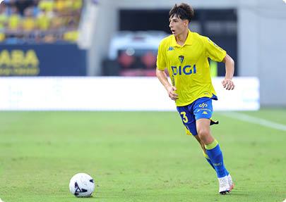 A person wearing a yellow sports jersey with the word DIGI is running on a soccer field with blurred spectators in the background. Descripci n generada con IA
