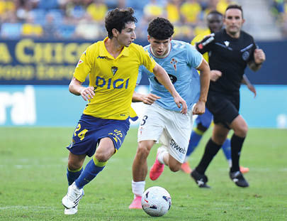Two adults in sports uniforms are actively competing for a soccer ball on a grass field with a referee running behind them. Descripci n generada con IA