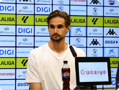 A person with short hair and a beard wearing a white t-shirt is sitting at a press conference table with microphones and branded bottles in front of a backdrop covered with various sponsor logos. Descripci n generada con IA