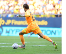 A person in an orange sports uniform and gloves is kicking a soccer ball on a grassy field with a blurred crowd and advertising banners in the background. Descripci n generada con IA