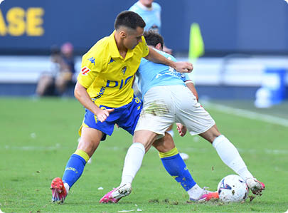 Two people in sports uniforms are competing for control of a soccer ball on a grassy field during a game. Descripci n generada con IA