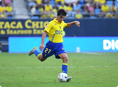 A person wearing a yellow and blue soccer uniform is kicking a soccer ball on a grass field during a game. Descripci n generada con IA
