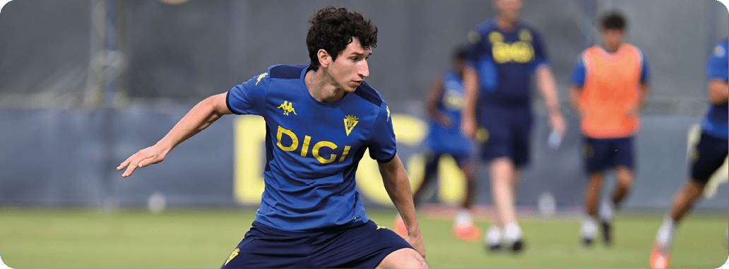 Una persona con uniforme deportivo azul y pantalones cortos est en posici n de juego sobre un campo de c sped con otras personas al fondo.  Descripci n generada con IA