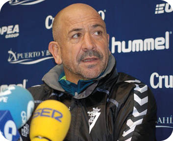 Una persona con chaqueta deportiva azul con detalles amarillos y rojos est se alando hacia un lado en un campo de c sped.  Descripci n generada con IA