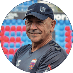 Persona adulta con gorra y camiseta deportiva posando en un estadio con gradas de colores rojo y azul desenfocadas al fondo. Descripci n generada con IA
