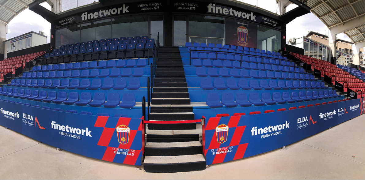 Gradas vac as de un estadio con asientos azules y rojos, con escaleras centrales y publicidad del club deportivo Eldense en los laterales. Descripci n generada con IA