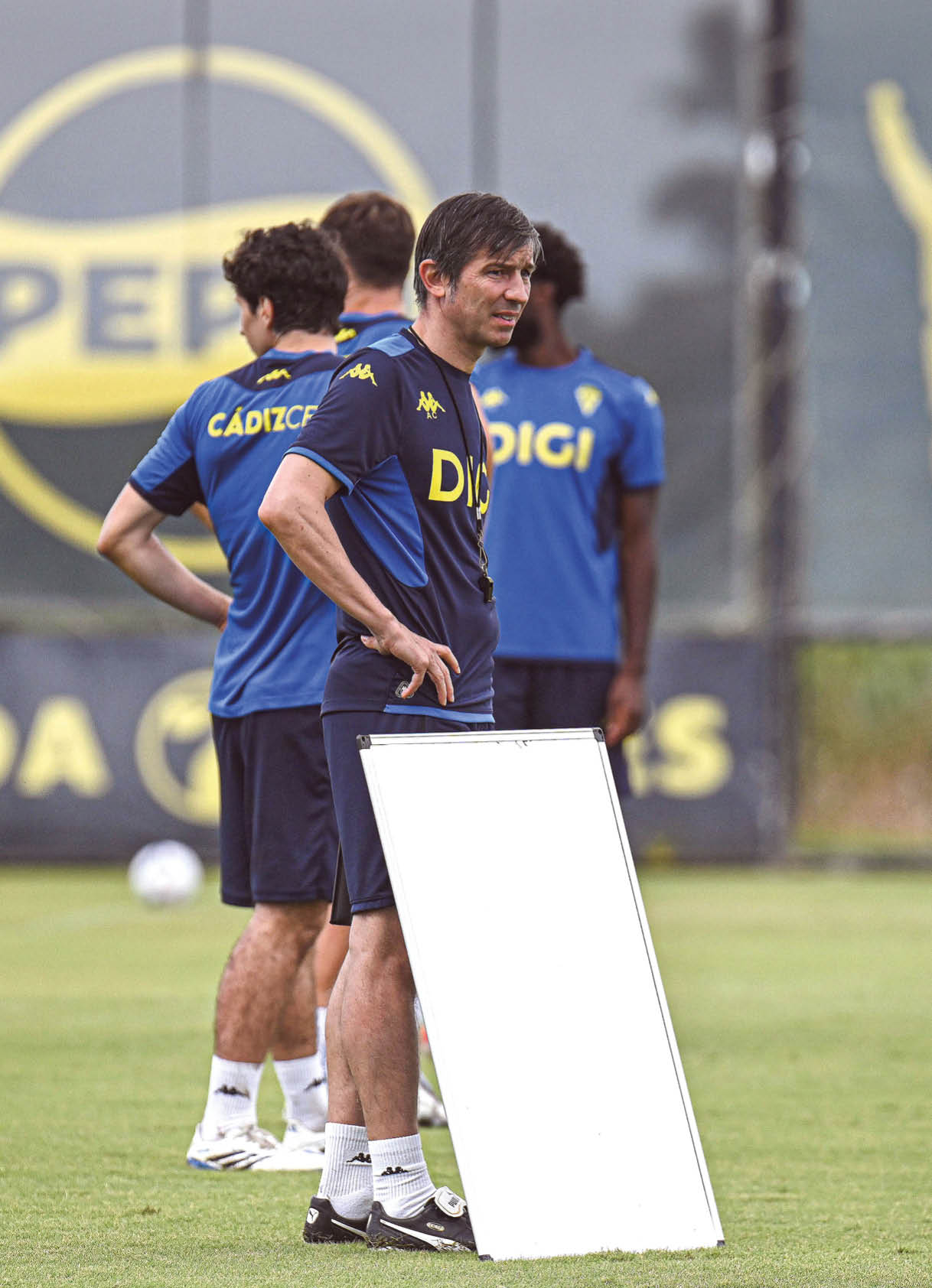 Una persona con ropa deportiva azul oscuro con el logo DIGI camina sobre un campo de c sped en un estadio con gradas vac as al fondo. Descripci n generada con IA