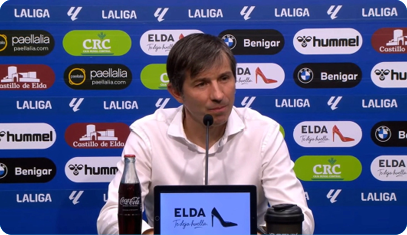 A person in a white shirt is sitting at a press conference table with a microphone, a bottle of Coca-Cola, and a coffee cup in front of a backdrop covered with various sponsor logos. Descripci n generada con IA