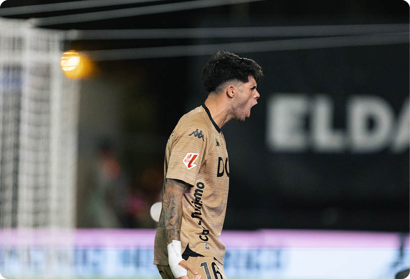 A person wearing a tan sports jersey with tattoos on their arm is shouting or celebrating near a soccer goalpost during a nighttime game. Descripci n generada con IA