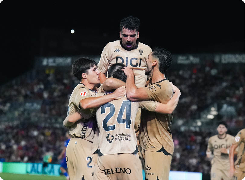 Four people wearing matching sports uniforms celebrate together on a field at night with a crowd in the background. Descripci n generada con IA