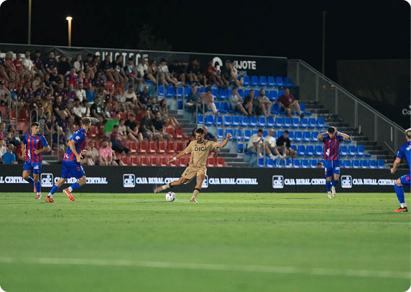 A person in a gold uniform is kicking a soccer ball on a field while several people in blue and red striped uniforms are positioned around them with spectators watching from the stands at night. Descripci n generada con IA