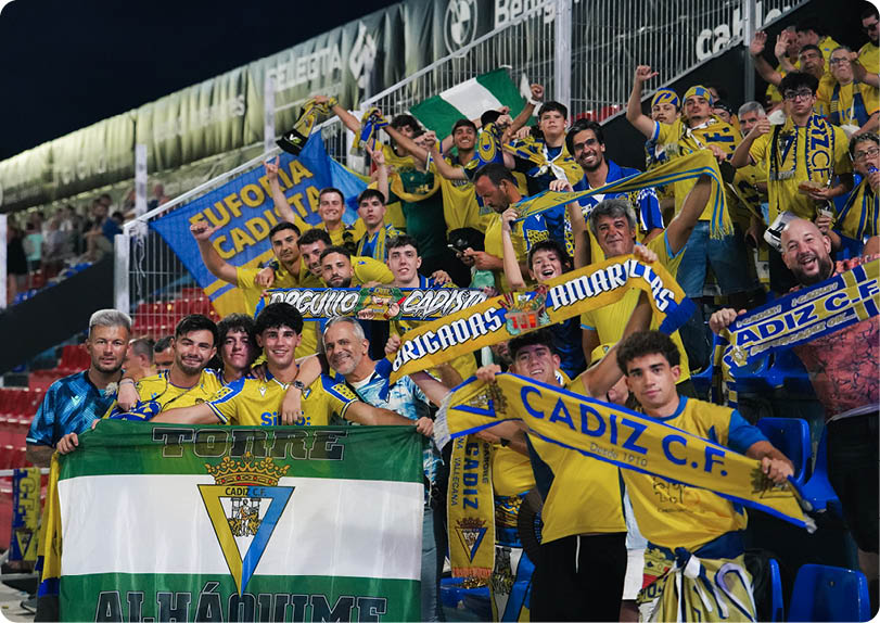 A group of enthusiastic people wearing yellow shirts and holding blue and yellow C diz C.F. scarves and flags are gathered in stadium seats, cheering and smiling. Descripci n generada con IA