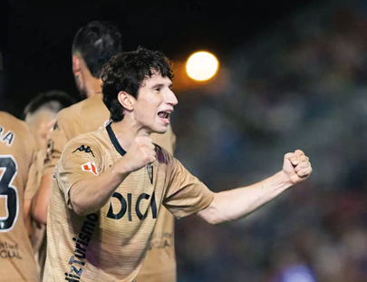 An adult wearing a gold sports jersey is celebrating with clenched fists and an open mouth, while other similarly dressed adults stand nearby in a stadium setting. Descripci n generada con IA