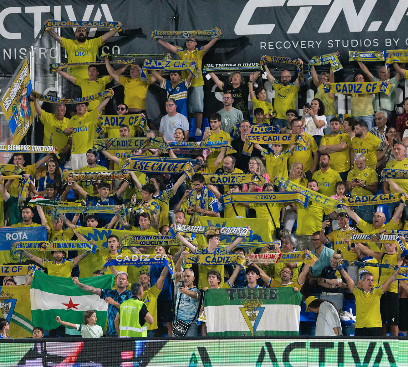 A large crowd of enthusiastic people wearing yellow shirts and holding blue and yellow scarves and flags is cheering in a stadium, supporting the C diz C.F. soccer team. Descripci n generada con IA