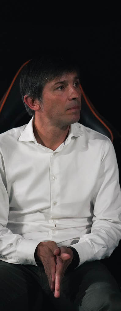 A person with short hair wearing a white button-up shirt is sitting with hands clasped, looking to the side against a dark background. Descripci n generada con IA