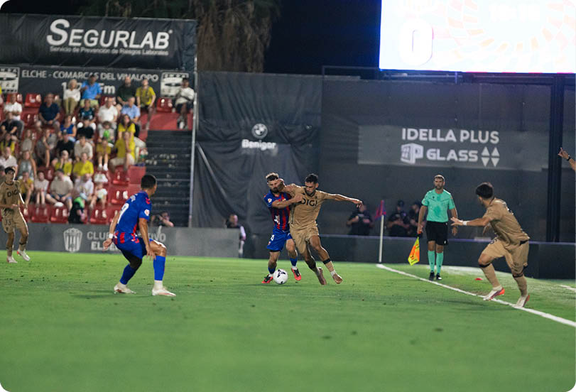 Two people in sports uniforms compete for a soccer ball on a field while a referee and other players watch near the sideline with spectators in the background. Descripci n generada con IA