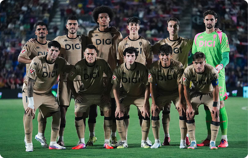 A group of eleven people wearing matching sports uniforms pose together on a soccer field with a crowd in the background. Descripci n generada con IA