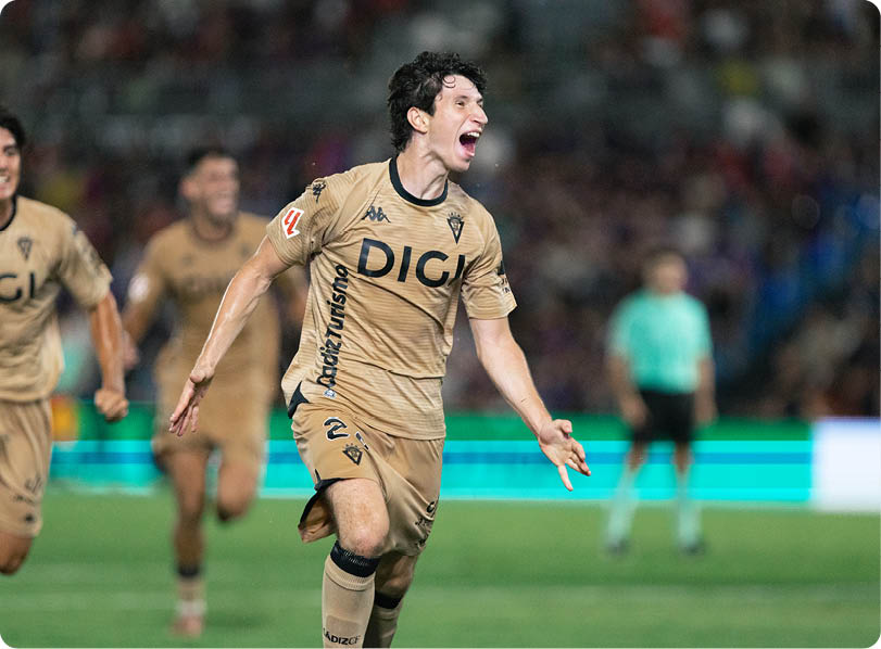 A person wearing a beige sports uniform is running on a field with an excited expression while other people in similar uniforms and a person in casual clothing are visible in the background. Descripci n generada con IA