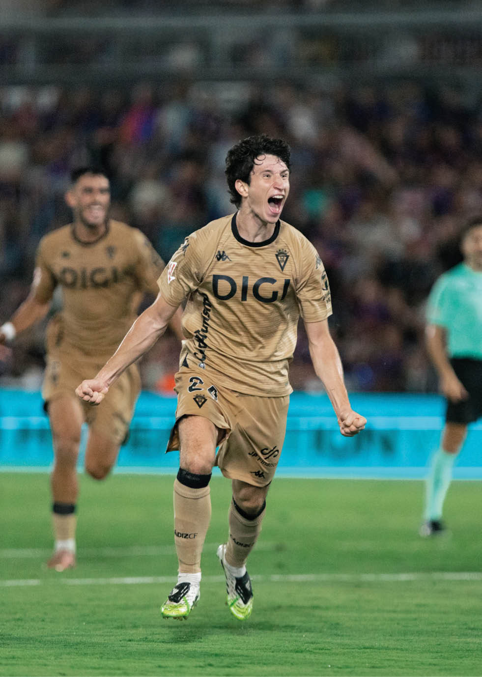 A person in a gold sports uniform is running on a soccer field with an excited expression while another person in a similar uniform runs behind. Descripci n generada con IA