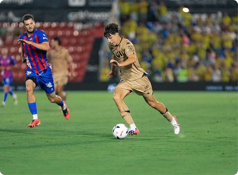Two adults in sports uniforms are playing soccer on a grass field with one person dribbling the ball while the other runs behind. Descripci n generada con IA